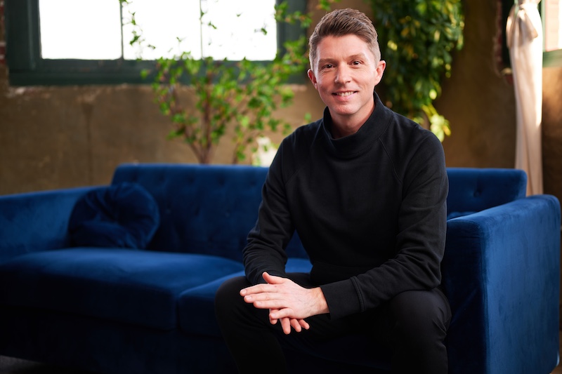 VP of Brand Strategy, Nathan Miller, sits on a blue velvet couch. He is dressed in all black with his hands in his lap. He smiles at the camera while discussing blogging for businesses.