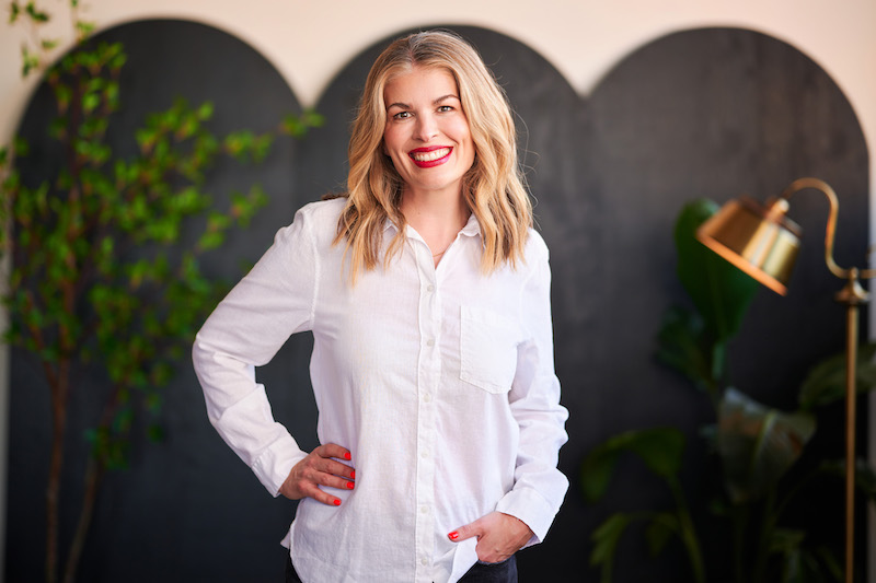 Formada founder, Meghan Kelly, wears a white button down shirt while standing in front of a black privacy screen, flanked by a plant to her left and a lamp to her right.