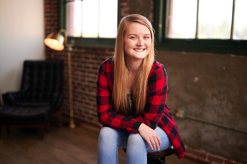 Natalie Nygren, wearing a black and red plaid shirt, sits on a chair, facing the camera and smiling.