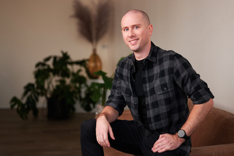 Formada VP of Design, David Kerr, is seated on a couch, wearing a gray and black plaid shirt and black jeans, smiling confidently at the camera.