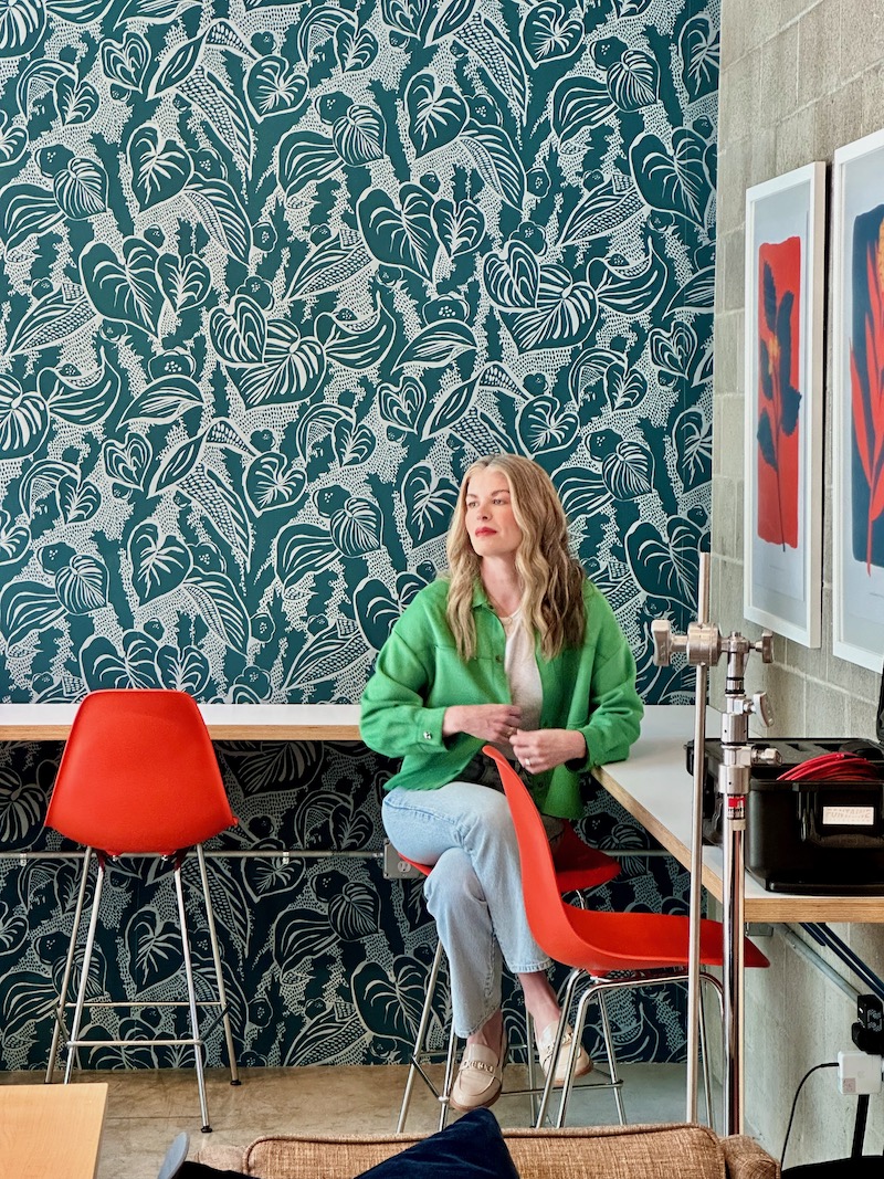 Formada Founder and CEO Meghan Kelly poses for a photo shot in front while sitting in a tall orange chair placed in front of a wall paper patterned wall.