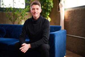 VP of Brand Strategy, Nathan Miller, is seated on a dark blue velvet couch, wearing all black clothes, discussing the benefits of a 4-day work week.