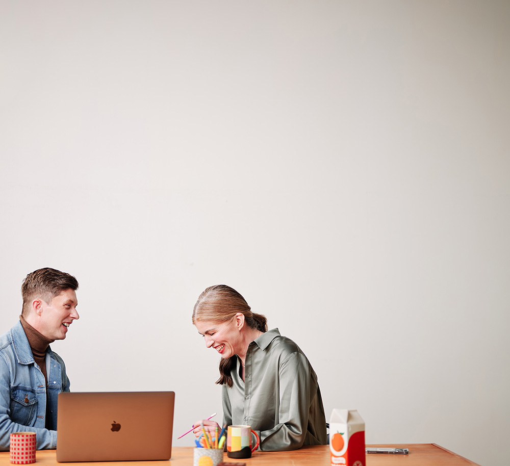 Formada's VP of Brand Strategy, Nathan Miller, is seated at a table behind a laptop, while he and Formada Founder, Meghan Kelly, laugh heartily together.
