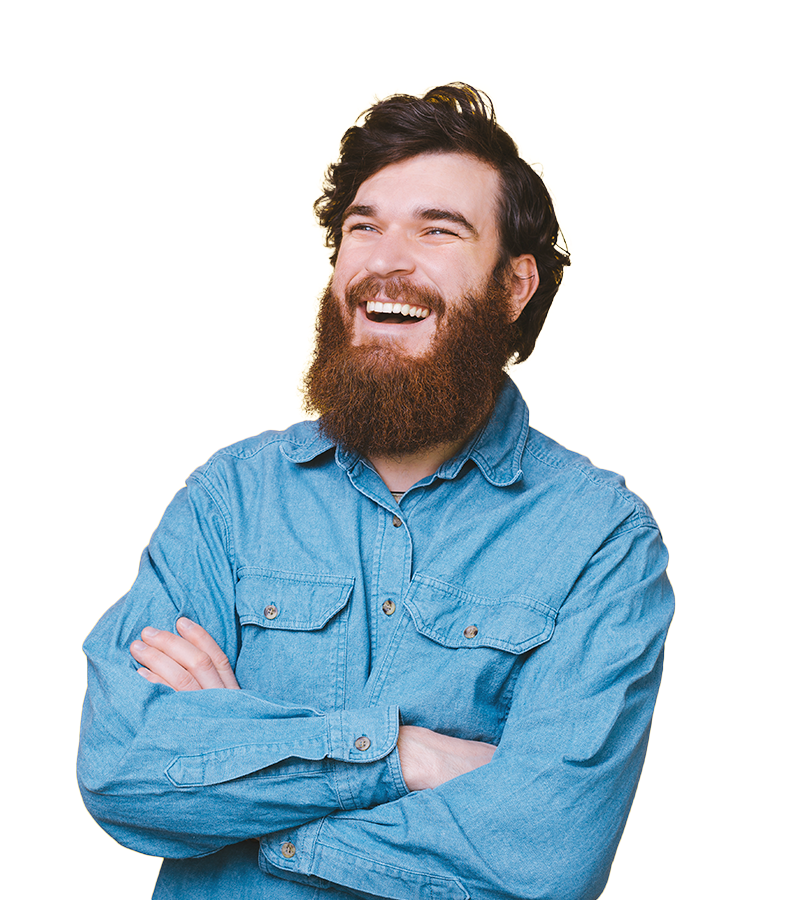 A young bearded man in a blue denim shirt, smiles and laughs while folding his arms and looking to the left.