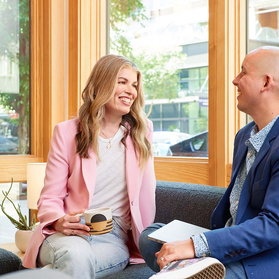 Formada's co-founders, Meghan Kelly and Garrett Jackson, smile at each other while sitting on a couch next to an office window