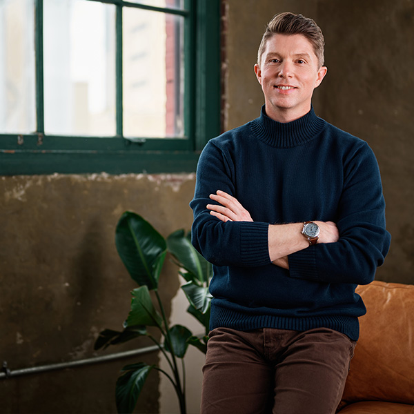 Formada VP of Brand Strategy, Nathan Miller, smiles while crossing his arms in front of an office couch and brick wall