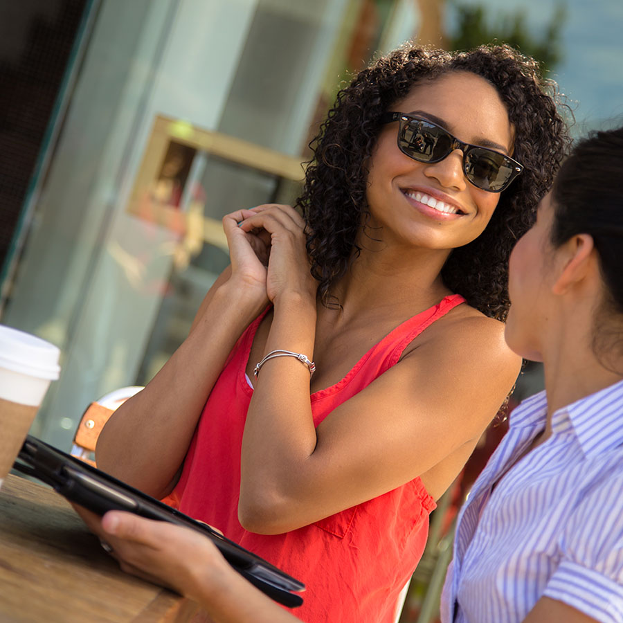 A woman smiles at her friend while sitting at a cafe as her friend hold a tablet