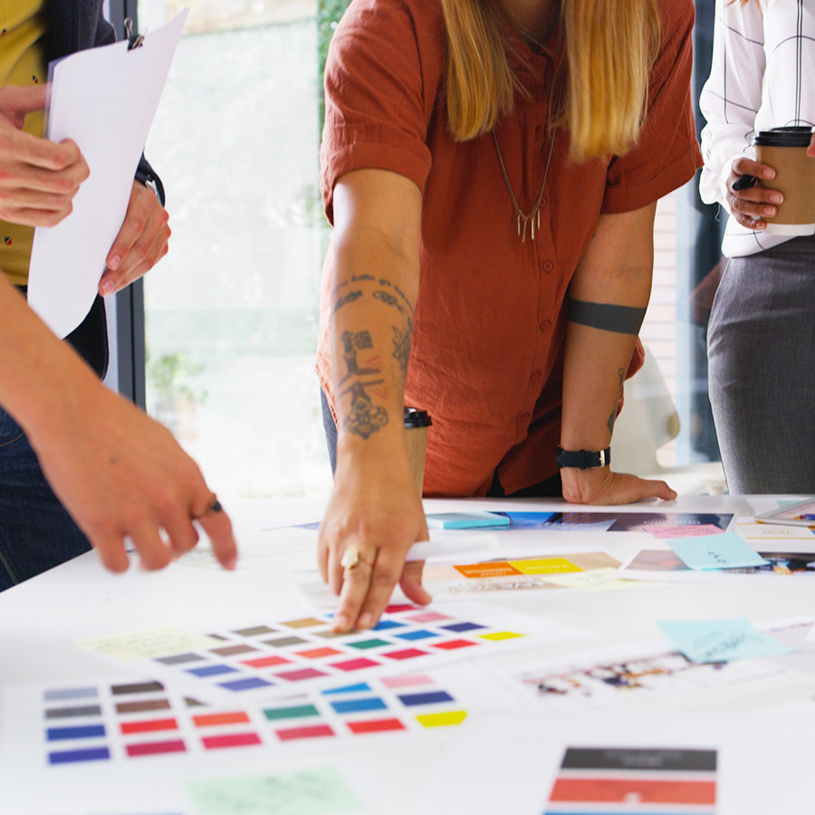 A team of four people, faces out of frame, surround a table covered in color swatches, discussing the merits of each.