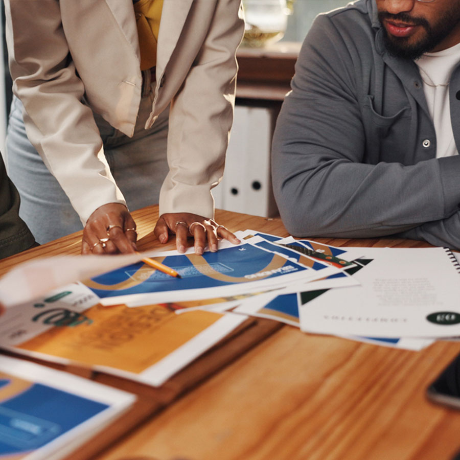 A woman holding a pencil, face obscured, goes over a printed presentation deck, while a seated man, face partially obscured, listens.