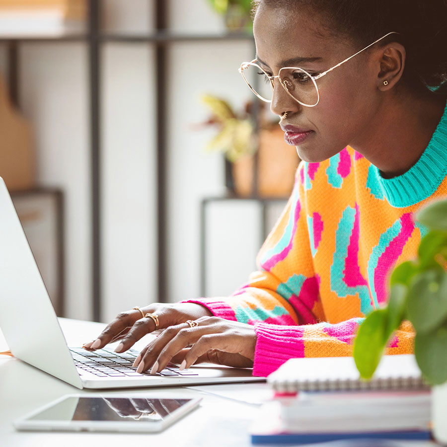 A young woman wearing glasses and a brightly patterned sweater is seated and typing on a laptop computer.