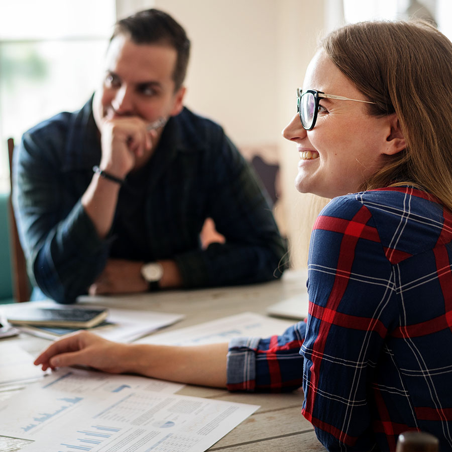 Two young professionals are seated at a table covered in printed charts, while smiling at a person speaking out of frame.